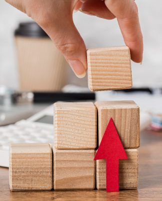 Una mano coloca un bloque de madera en una pila junto a una flecha roja, con una calculadora y una taza de café en el fondo.