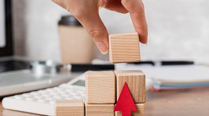 Una mano coloca un bloque de madera en una pila junto a una flecha roja, con una calculadora y una taza de café en el fondo.